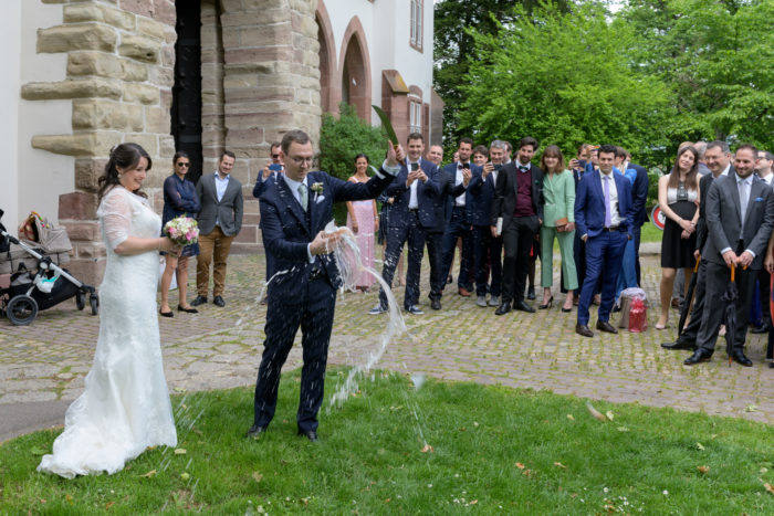 Bräutigam sabriert den Champagner vor den Gästen im Freien. Braut steht etwas abseits und schaut zu.