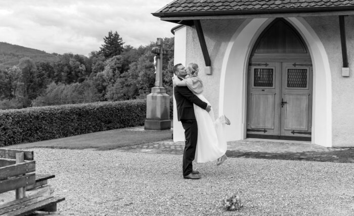 Hochzeitspaar küsst sich vor einer Kapelle. Bräutigam hat Braut hochgehoben.