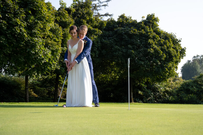 Brautpaar steht auf einem Golfplatz bei der Puttinganlage und schlägt einen Golfball Richtung Loch.