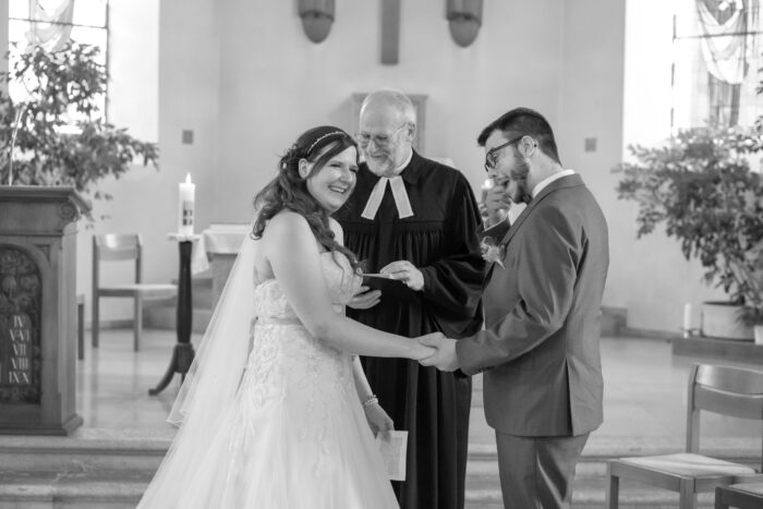 Brautpaar steht vor dem reformierten Pfarrer. Die Braut lächelt, der Bräutigam wischt sich die Freudenträne aus dem Auge und der Pfarrer schmunzelt.